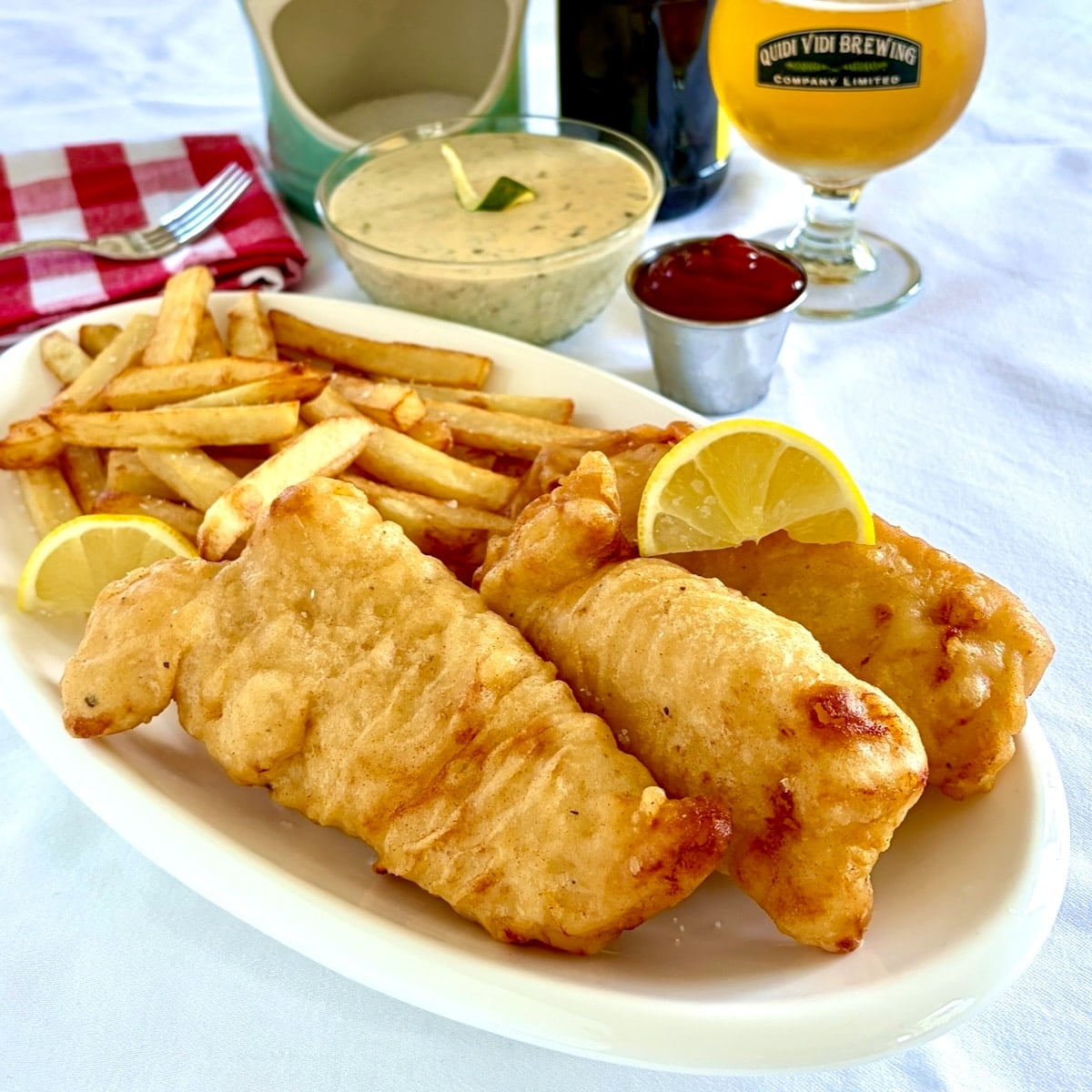 Fish and chips shown with tartar sauce and a glass of beer in the background.