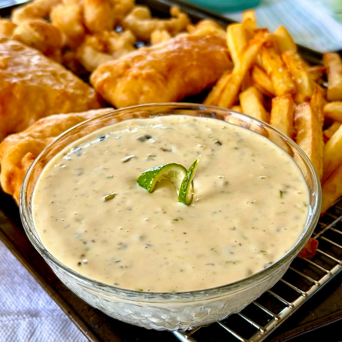 Lime Caper Tartar Sauce photo of sauce in a clear glass serving bowl with fish and chips cooling on a wire rack in the background.